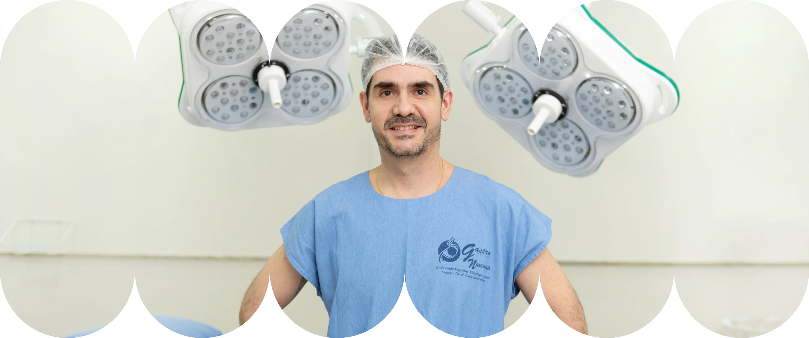 O Dr. Renan sorrindo e olhando para a câmera, vestindo um uniforme cirúrgico azul (scrubs) e touca, em pé em uma sala de cirurgia. Duas luminárias cirúrgicas modernas estão penduradas acima de uma maca cirúrgica.
