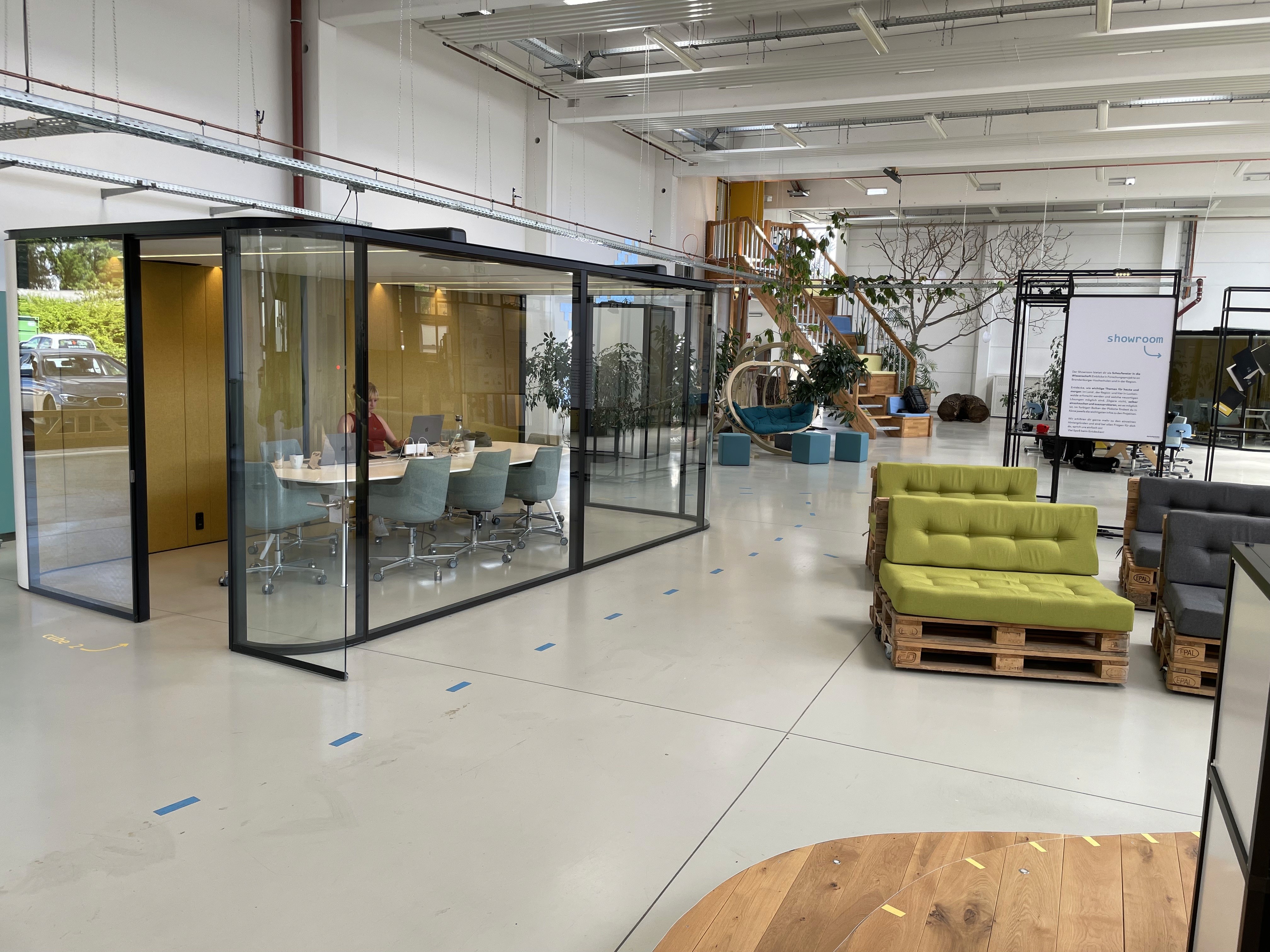 Foto aus dem Corworkingspace Luckenwalde. Großer Space mit Glascubes für Besprechungen, Sitzmöglichkeiten, eine Bühne mit Rednerpult, Ausstellungsaufsteller