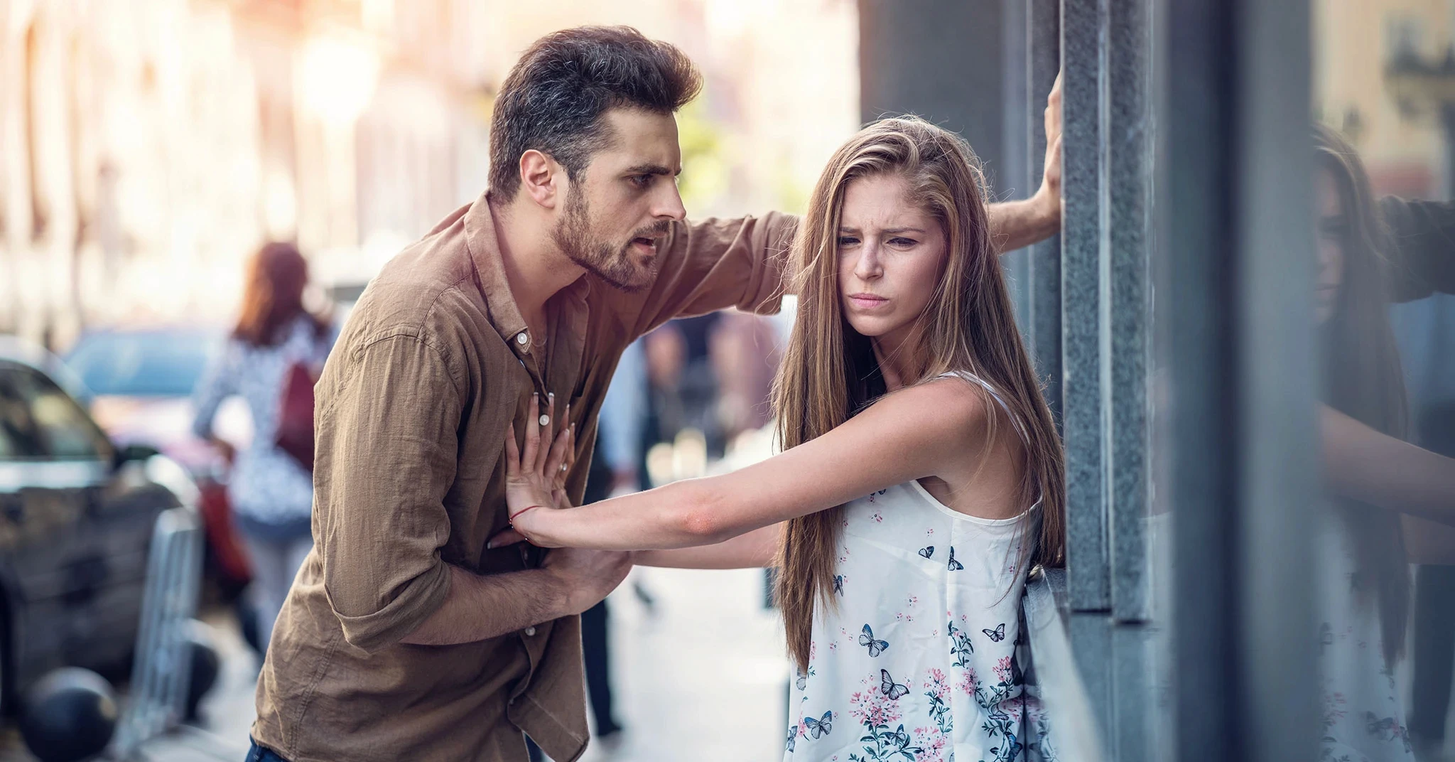 En man i en brun jacka står och diskuterar med en irriterad kvinna som lutar sig mot en vägg på en livlig gata.