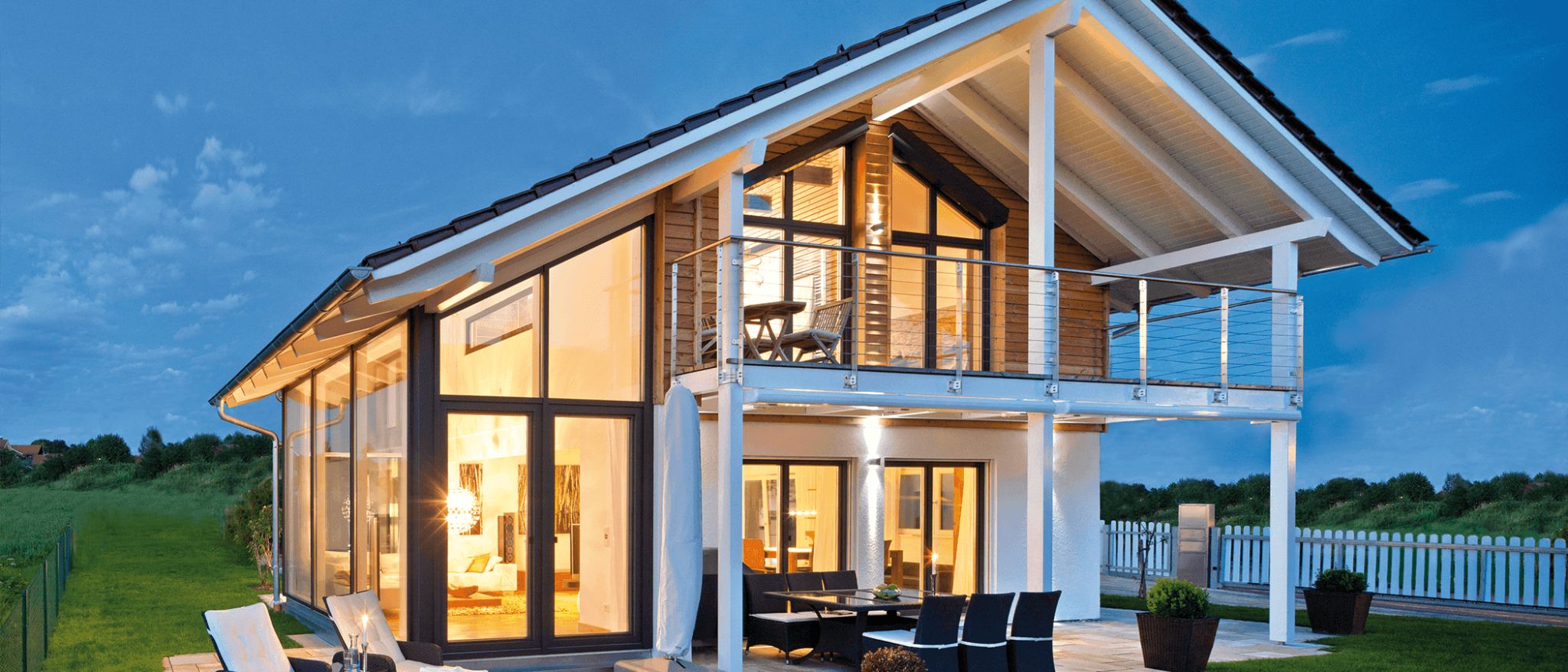 Modernes Haus mit großzügigen Glasfronten, Balkon mit Edelstahlgeländer und möblierter Terrasse im Abendlicht.