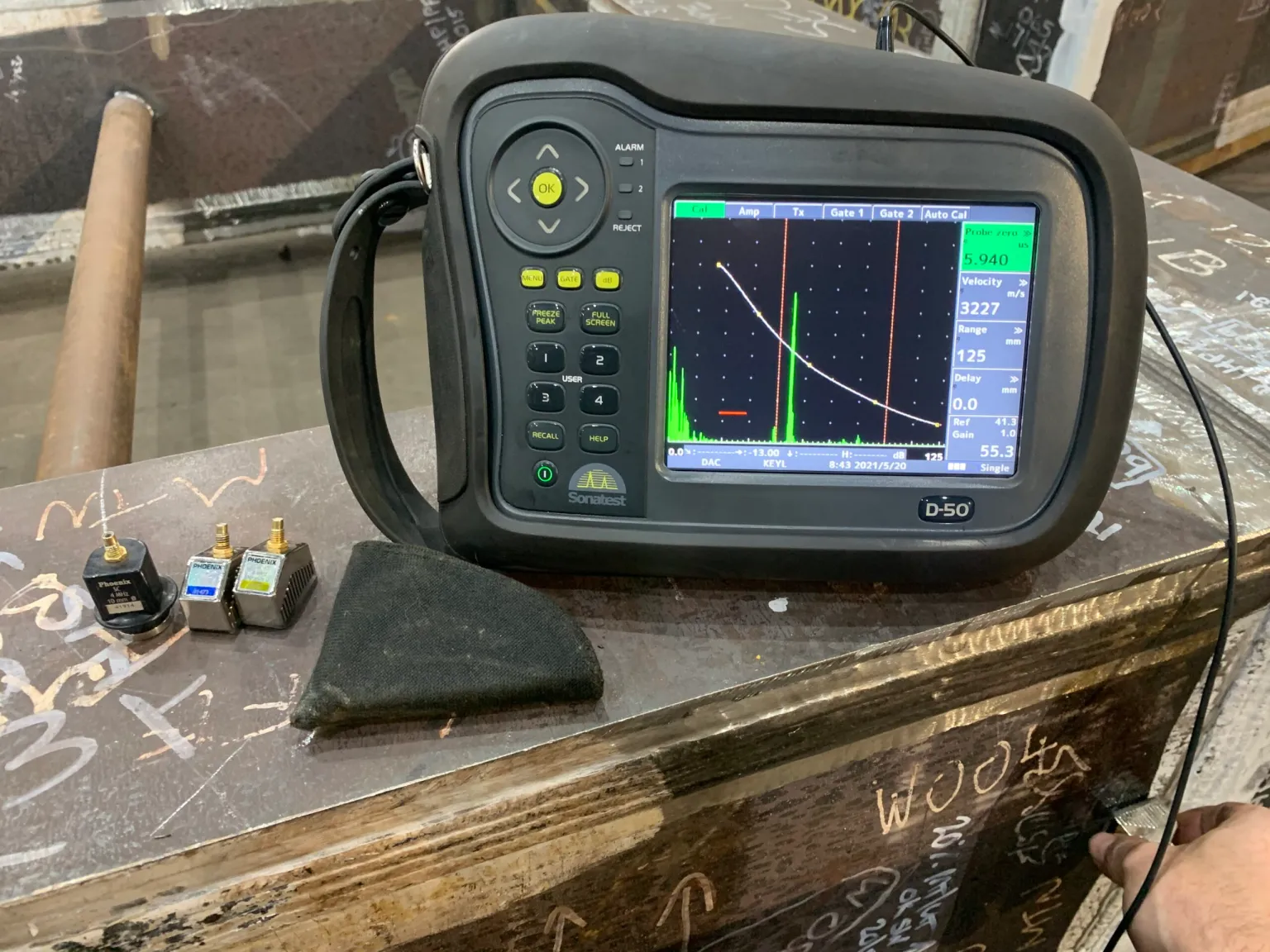 Ultrasonic testing device displaying a graph on its screen, placed on a marked metal surface. Nearby are three small components and a carrying case.