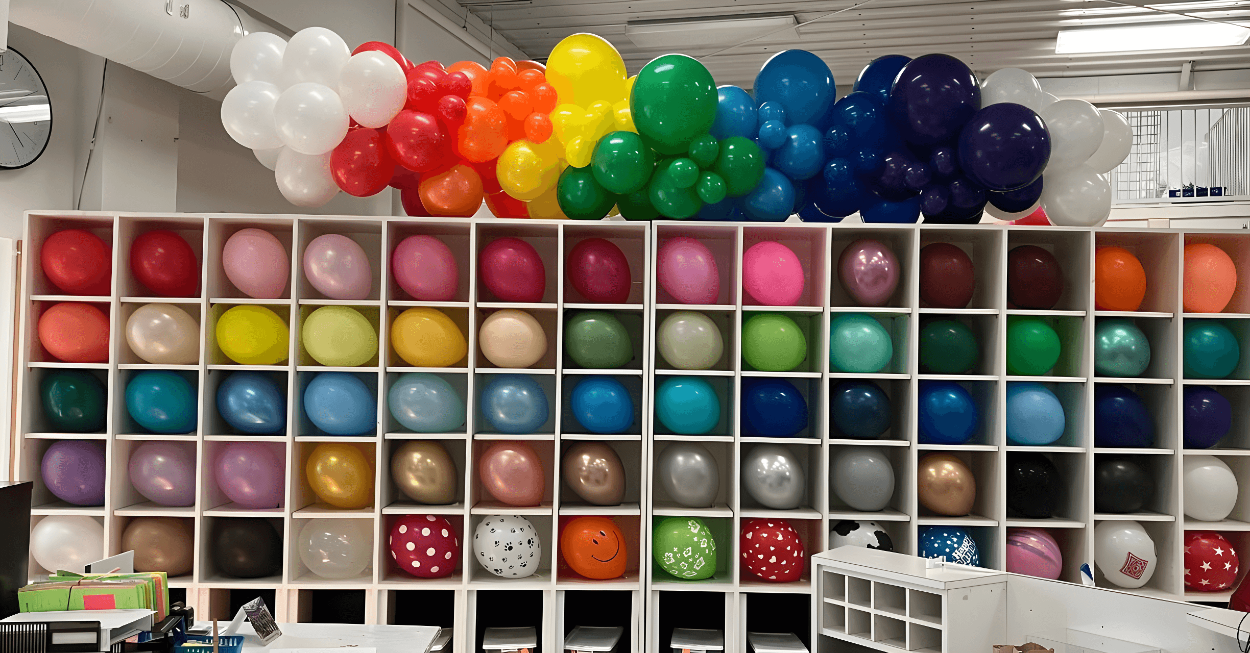 A vibrant array of colorful balloons is arranged artfully in and above a grid of cubbyholes, showcasing a spectrum from red to violet, in a spacious, well-organized room.