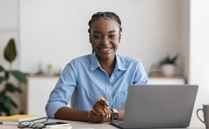 A imagem mostra uma mulher jovem sorridente, de pele escura, sentada em frente a um laptop em uma mesa. Ela está usando uma camisa azul clara e tem as mãos apoiadas na mesa, parecendo estar em um ambiente de trabalho. Ao lado do laptop, há óculos, papéis e uma caneca, indicando que ela está em um momento de concentração ou produtividade. O fundo está desfocado, sugerindo um espaço de escritório ou ambiente doméstico.