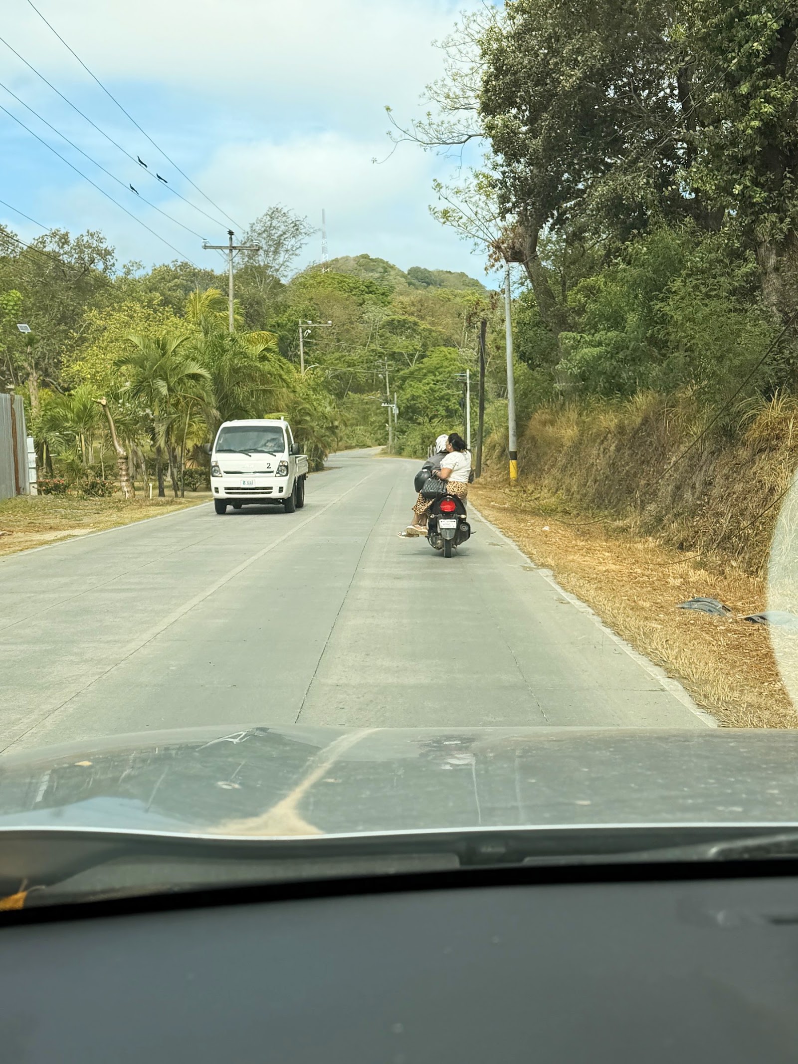 A narrow concrete road across Roatán with a scooter carrying two riders and a small truck approaching