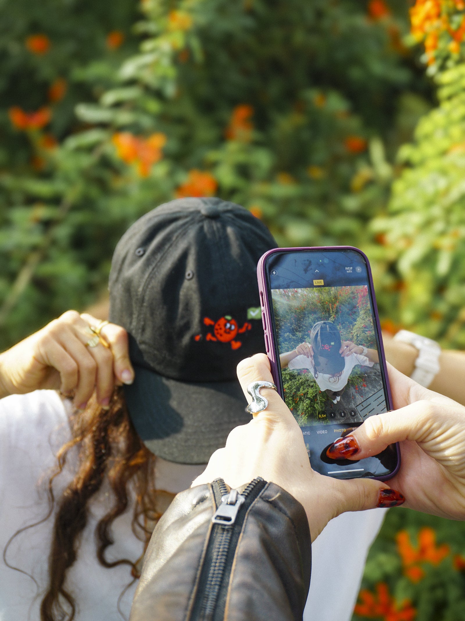 Person taking a photo of another person wearing a black cap with an orange fruit design using a phone.