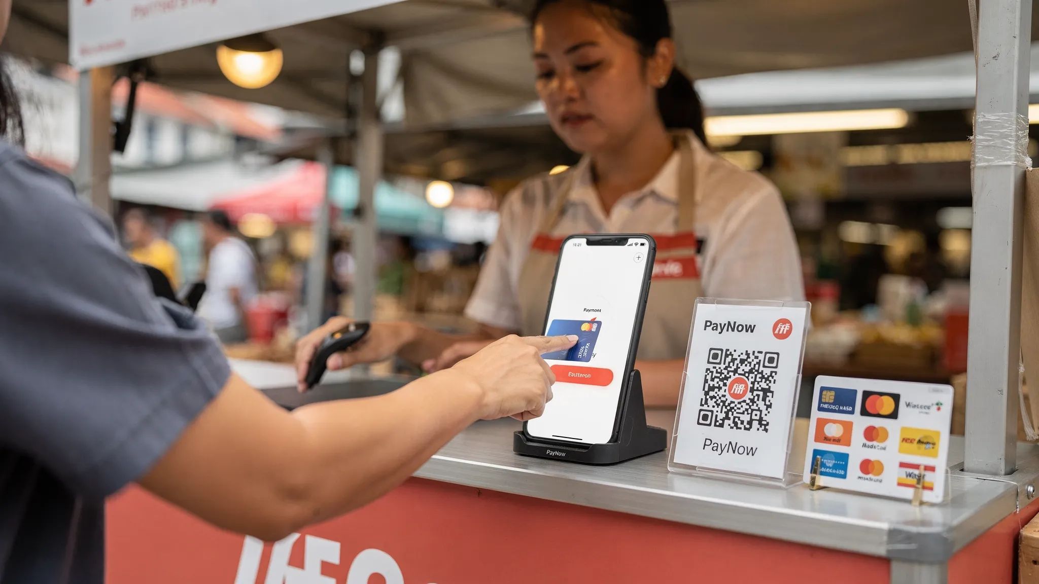 A Singapore pop-up stall checkout counter where a customer taps a contactless card on a merchant smartphone; the phone screen is upright and facing the merchant, with a PayNow QR sign and card logos nearby.