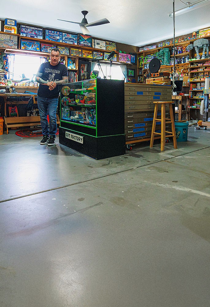 Artist Dirty Donny Gillies standing in his brightly lit studio filled with vintage toys, art supplies, and colorful collectibles, surrounded by shelves packed with models, posters, and pinball machines.