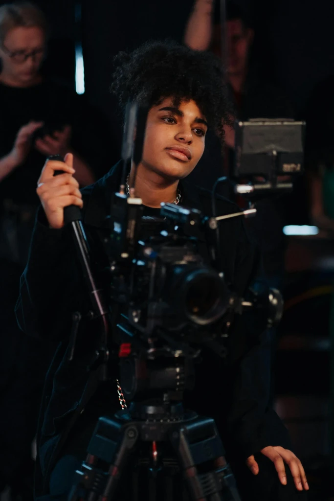 Focused woman with curly hair operating a professional cinema camera on a dark film set.