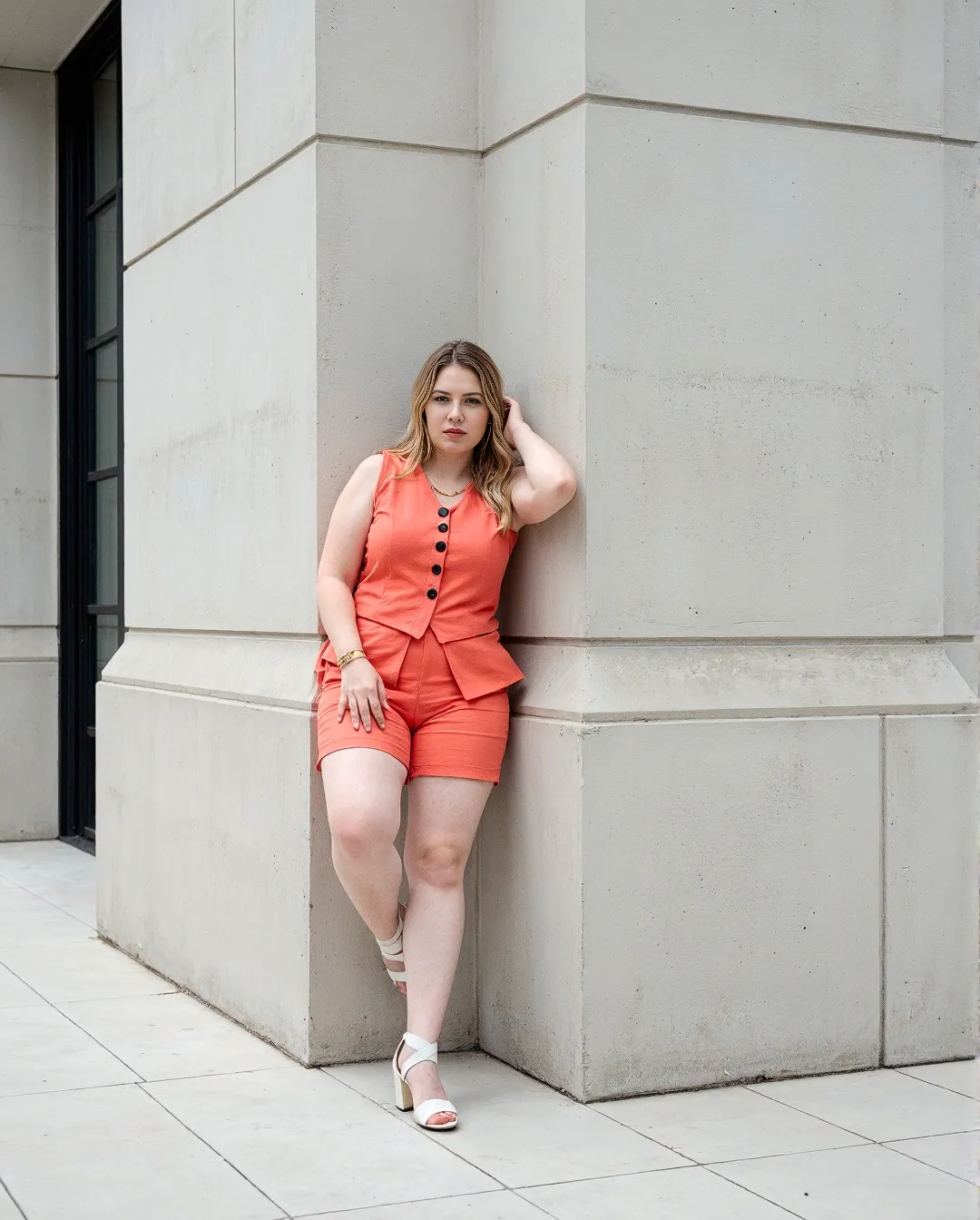 Stylish coral two-piece outfit featuring button-front vest and tailored shorts, posed against minimalist concrete architecture