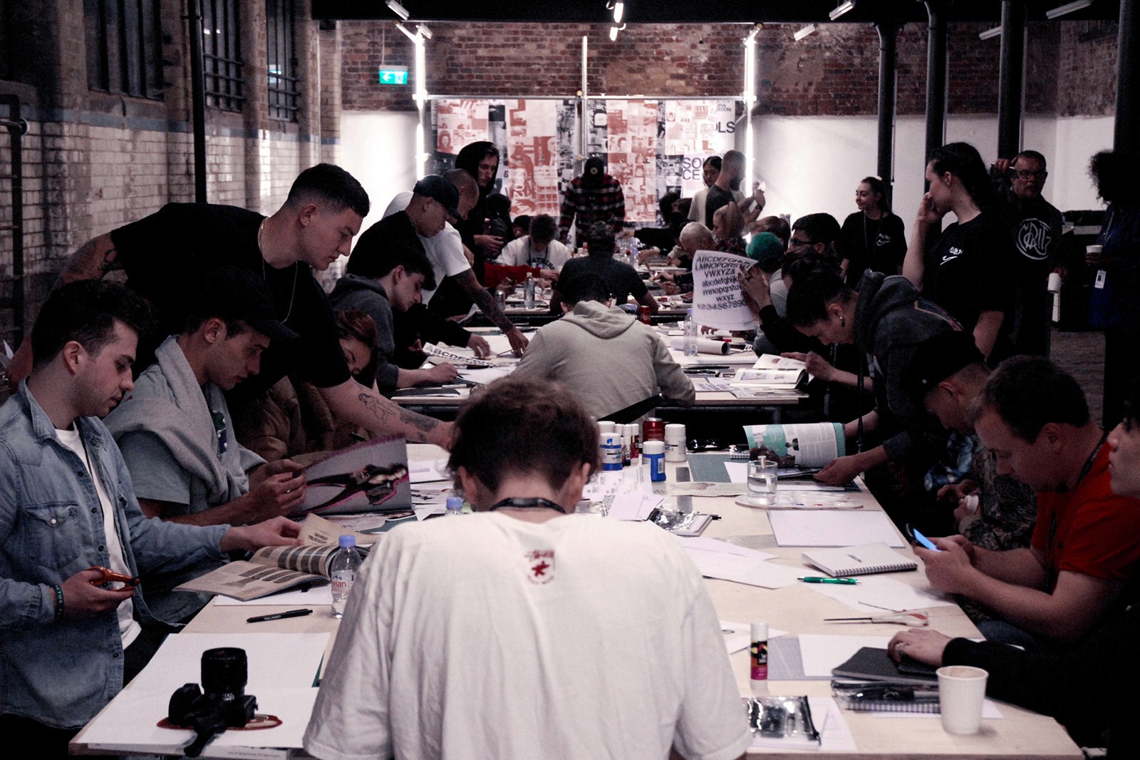A large group of focused artists, seated around a long table covered with art supplies, collaborate intensely in a creatively vibrant workshop within an industrial-style room.
