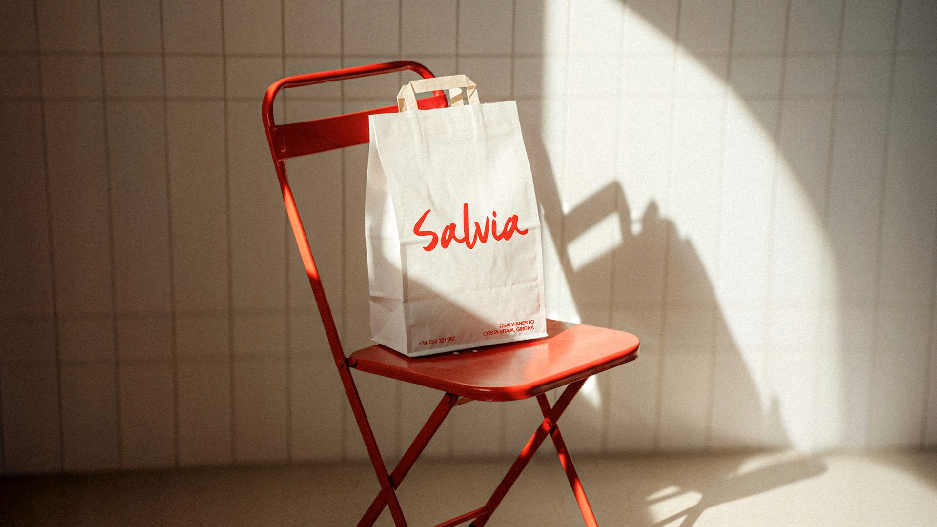A red metal folding chair is placed in front of a sunlit tiled wall, with a white paper bag that has "Salvia" written in red lettering resting on the seat.