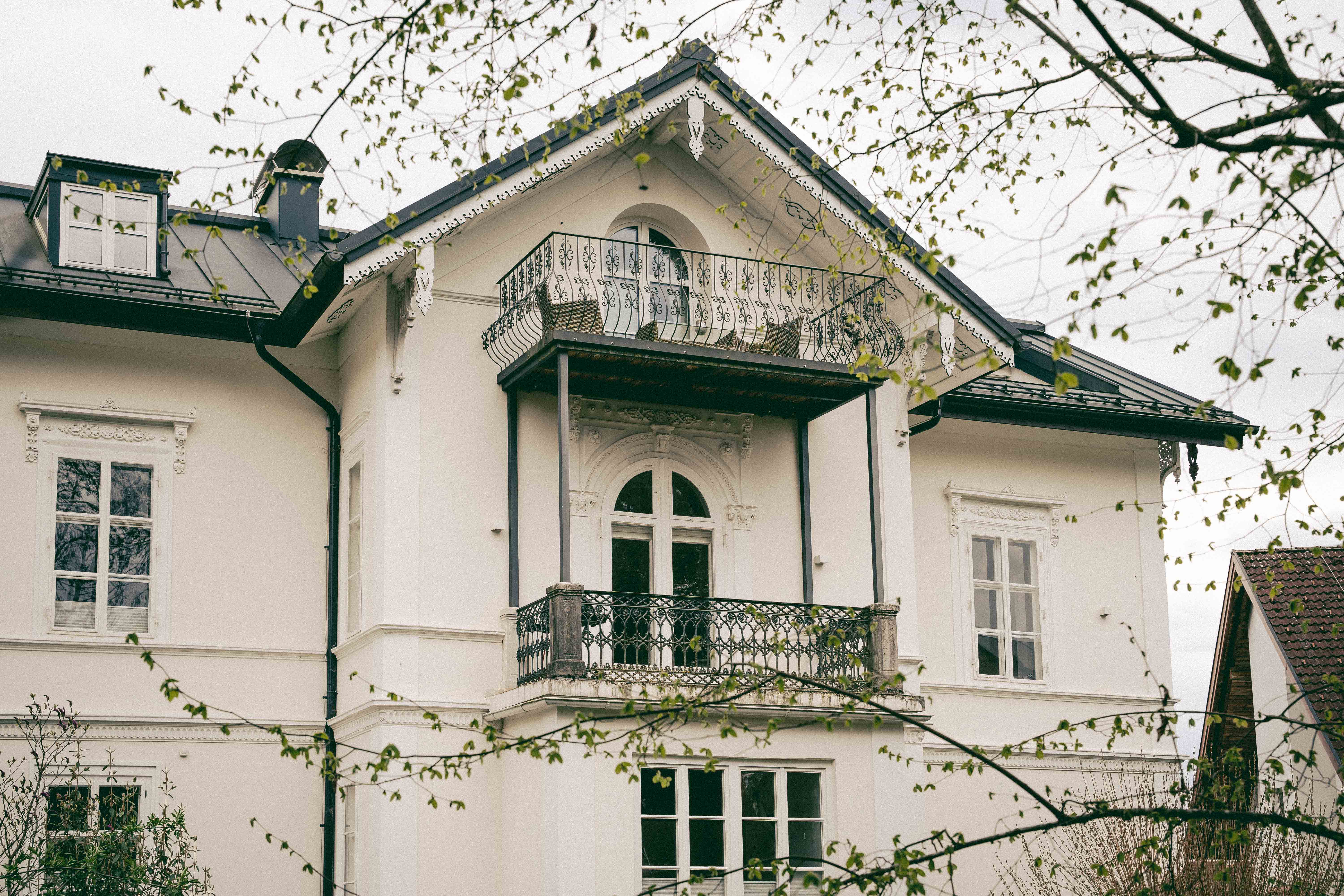 Weißes Haus mit Balkon, schwarzem Dach und Sprossenfenstern, umgeben von Ästen und Blättern.