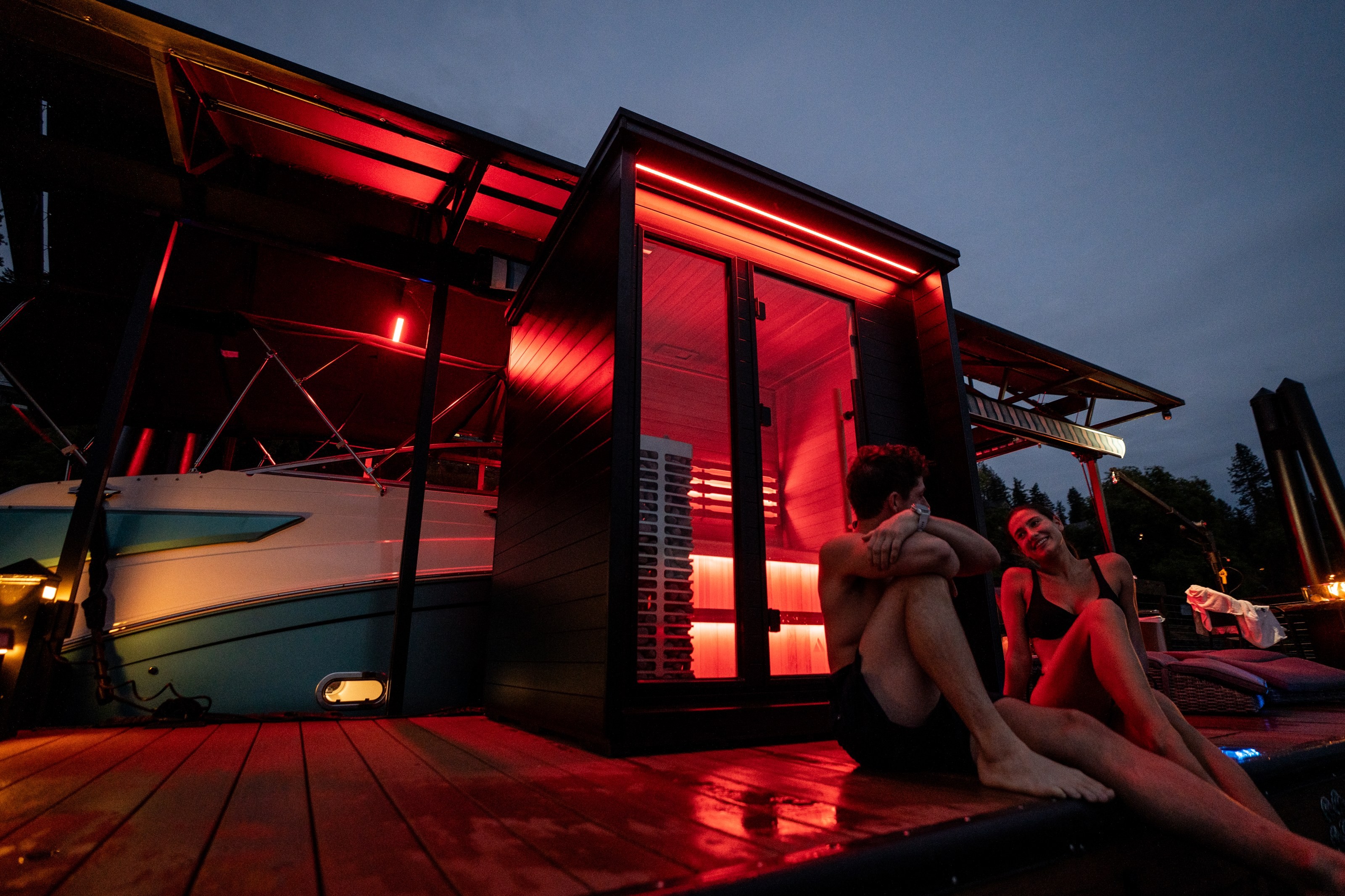 Outdoor sauna glowing at dusk beside a lake, with two people resting on the dock.