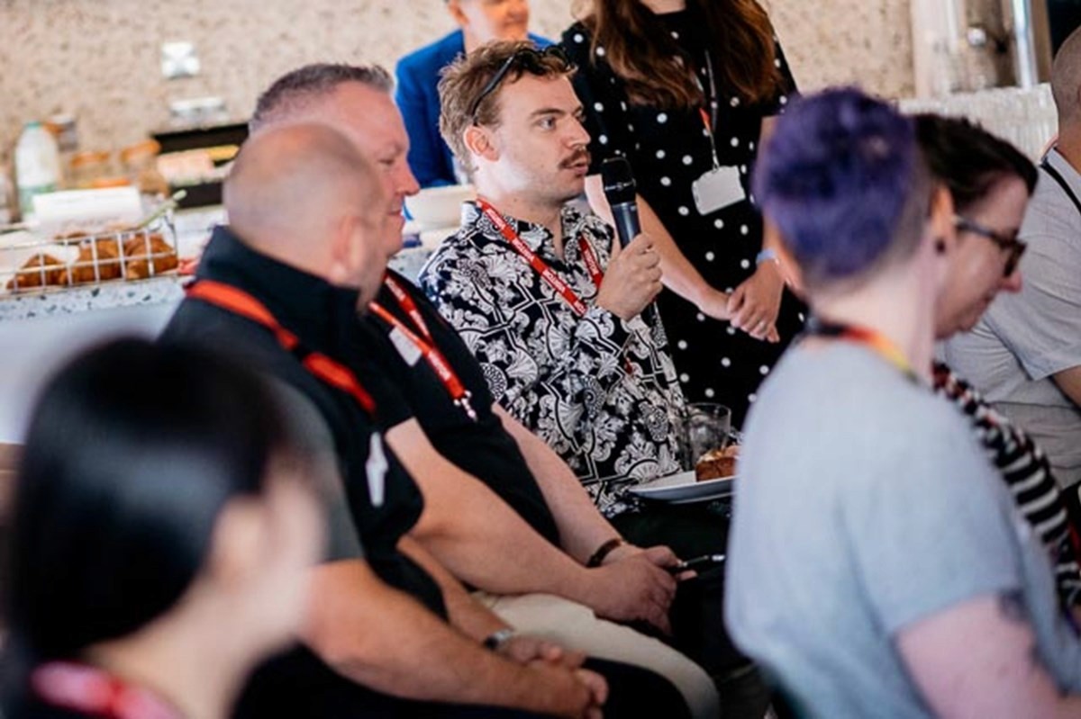 A group of people seated closely together indoors during a discussion or Q&A session. One person in the centre is holding a microphone while speaking, wearing a patterned top and an event lanyard. Others nearby are listening attentively, some with hands folded or resting on their laps. The setting appears informal and social, with refreshments visible in the background, suggesting a community or workplace event.