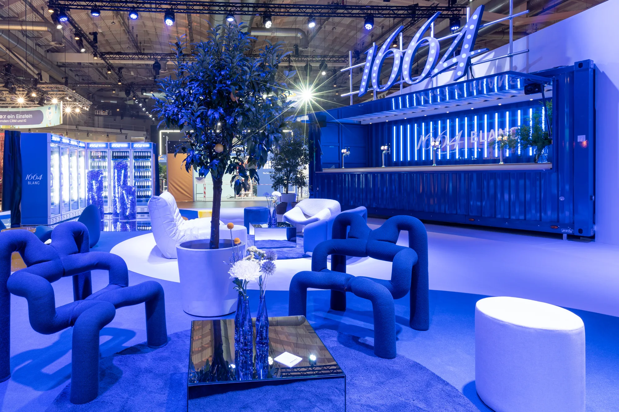 1664 Blanc trade fair lounge in monochrome blue, with sculptural tubular armchairs, a central olive tree planter and an illuminated bar in the background.