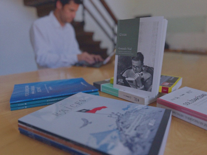 Revistas y libros de IdeaPaís se exponen sobre una mesa, mientras atrás, fuera de foco, una persona trabaja en su computador.