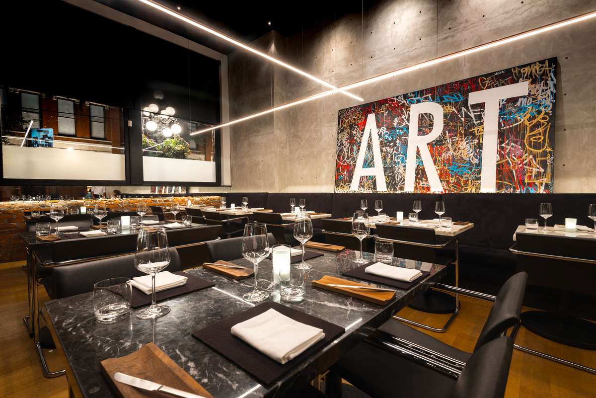 A modern restaurant interior with dark tables set with white plates and wine glasses in front of a large colorful mural.