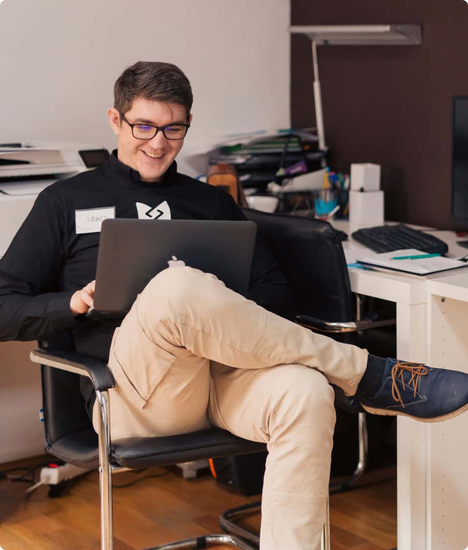 Smiling emplyee in glasses sitting in an office chair, working on a laptop, exuding a relaxed and happy demeanor.