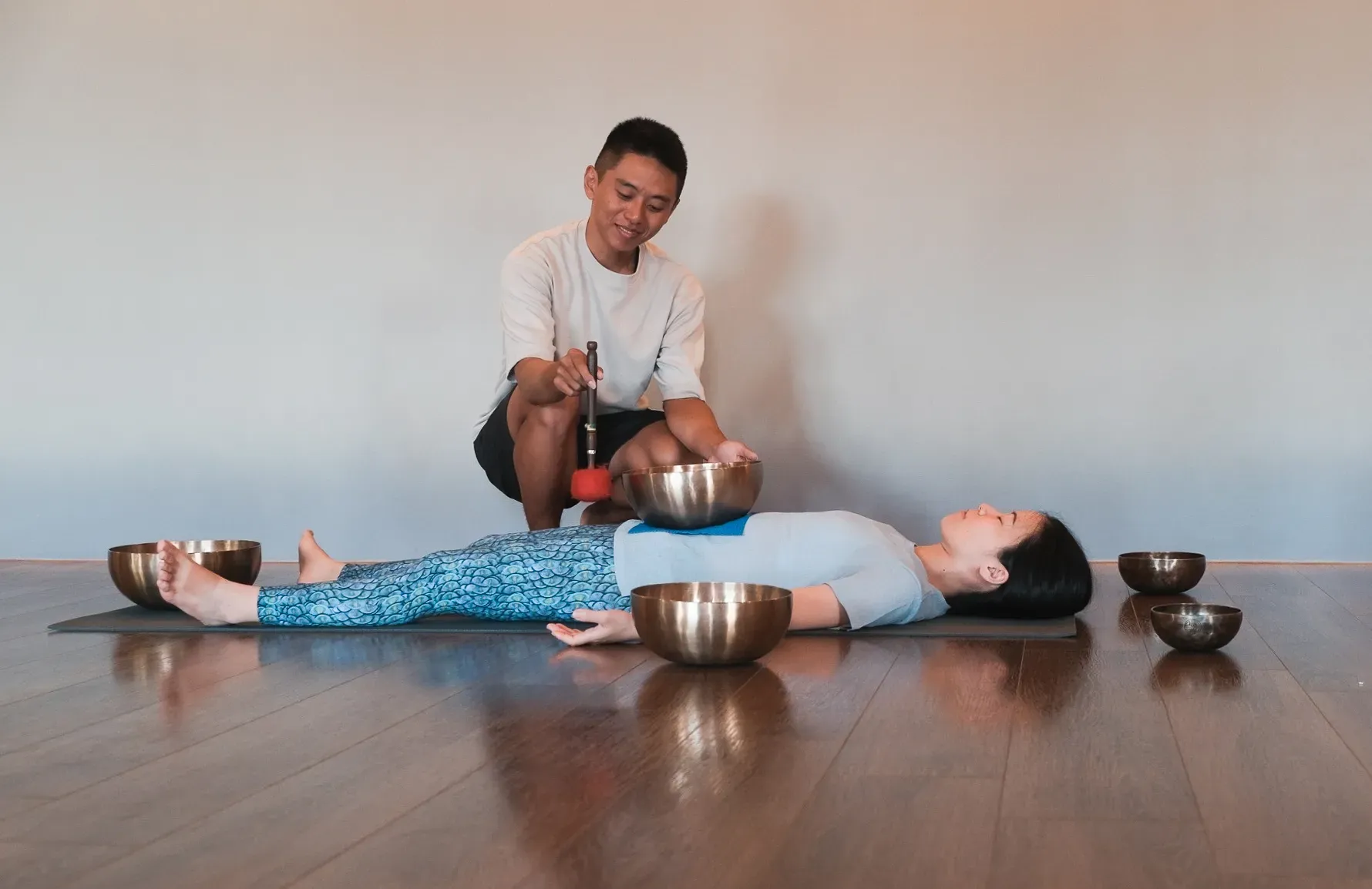 男引導師蹲在台北的木地板上,用木槌敲打頌缽;一名女子躺在他身旁,軀幹上擺放著另一個頌缽。