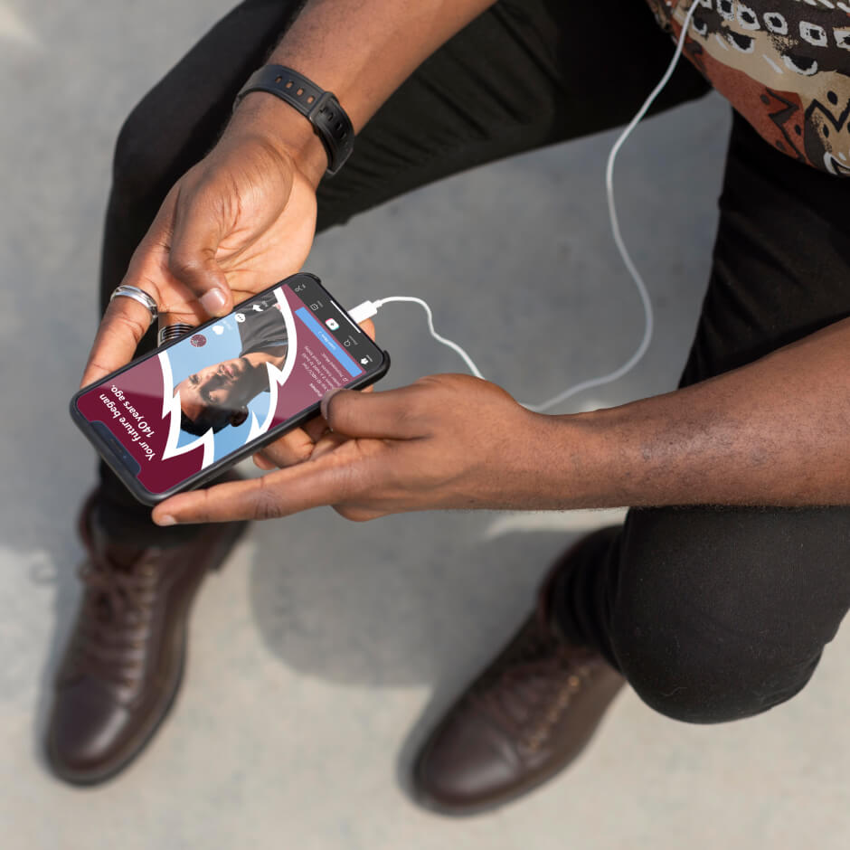 A person holding a smartphone with earphones plugged in, viewing a podcast episode on the screen. The person is wearing dark pants, brown boots, and a patterned shirt.