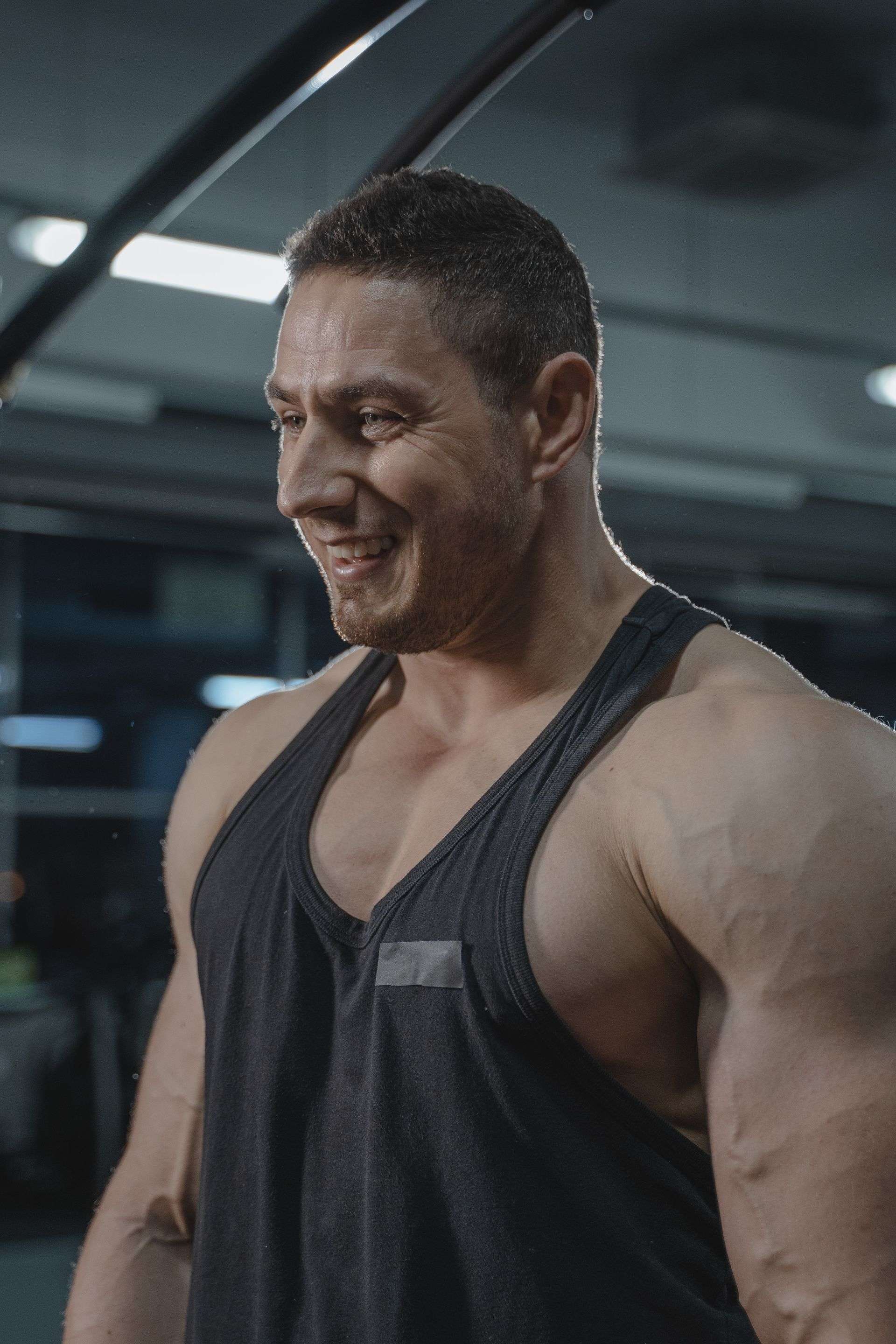 Fit man in a black tank top smiling during workout in gym. Strong and muscular build captured indoors.