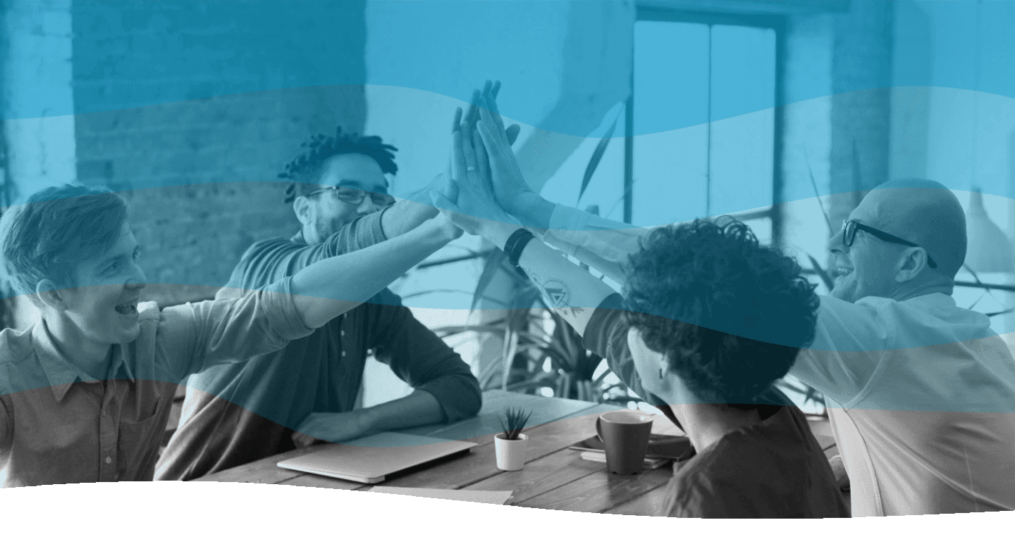 Four people sitting at a wooden table in a bright, modern office, celebrating with a high-five; laptops, papers, and a small plant adorn the workspace.