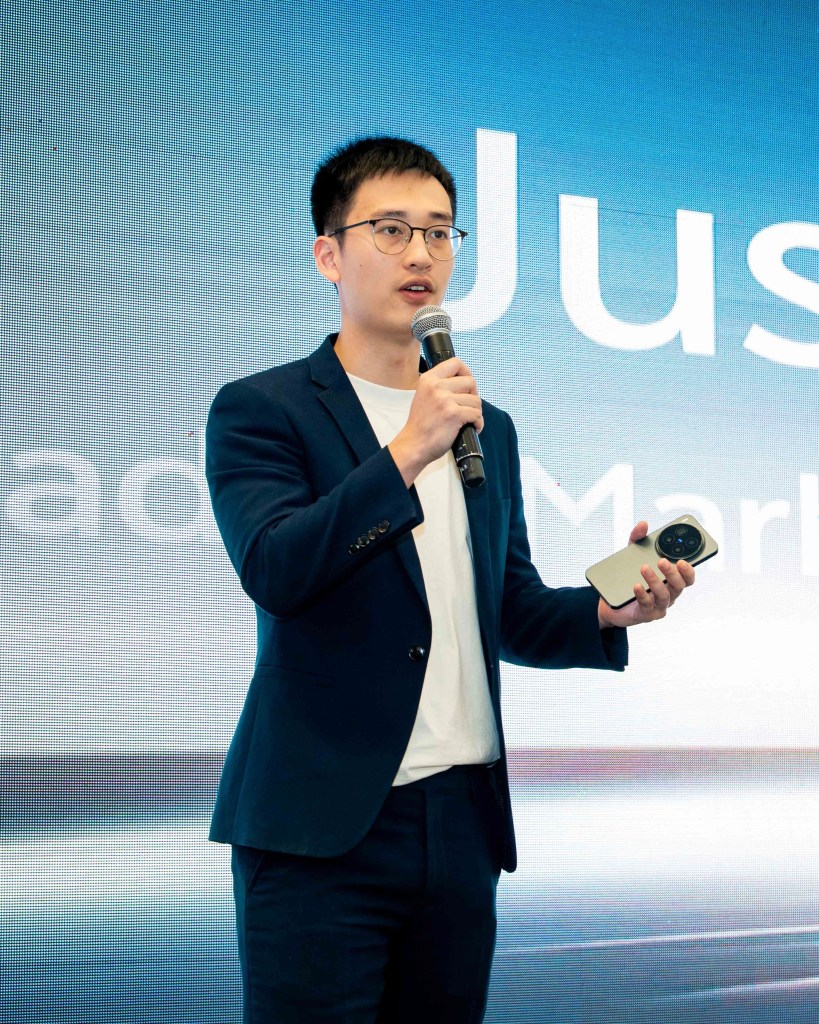 A person in a suit holding a smartphone while speaking at an event, with a screen displaying text behind them.