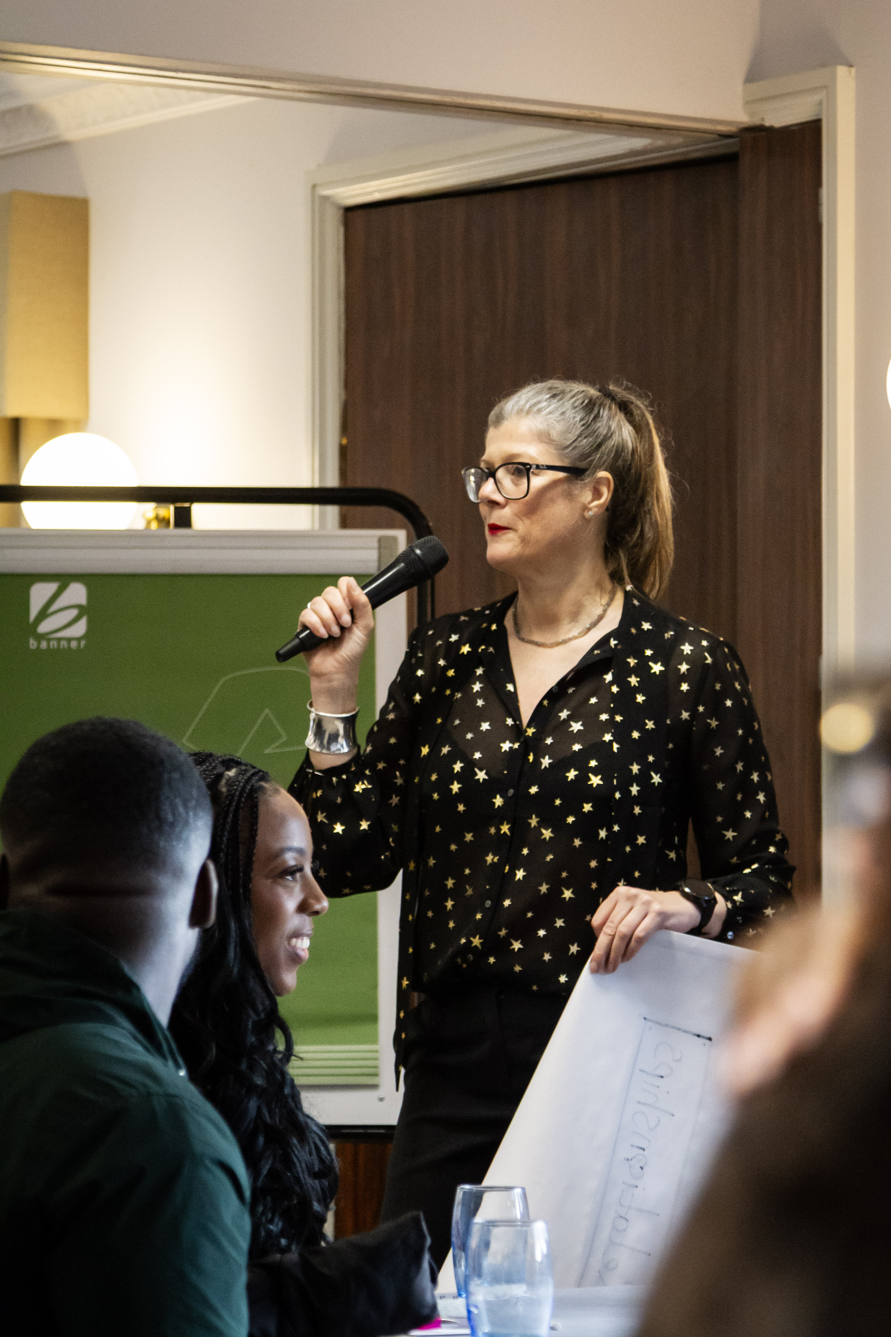 Woman with glasses and a black polka dot shirt speaks into a microphone while holding a blueprint.