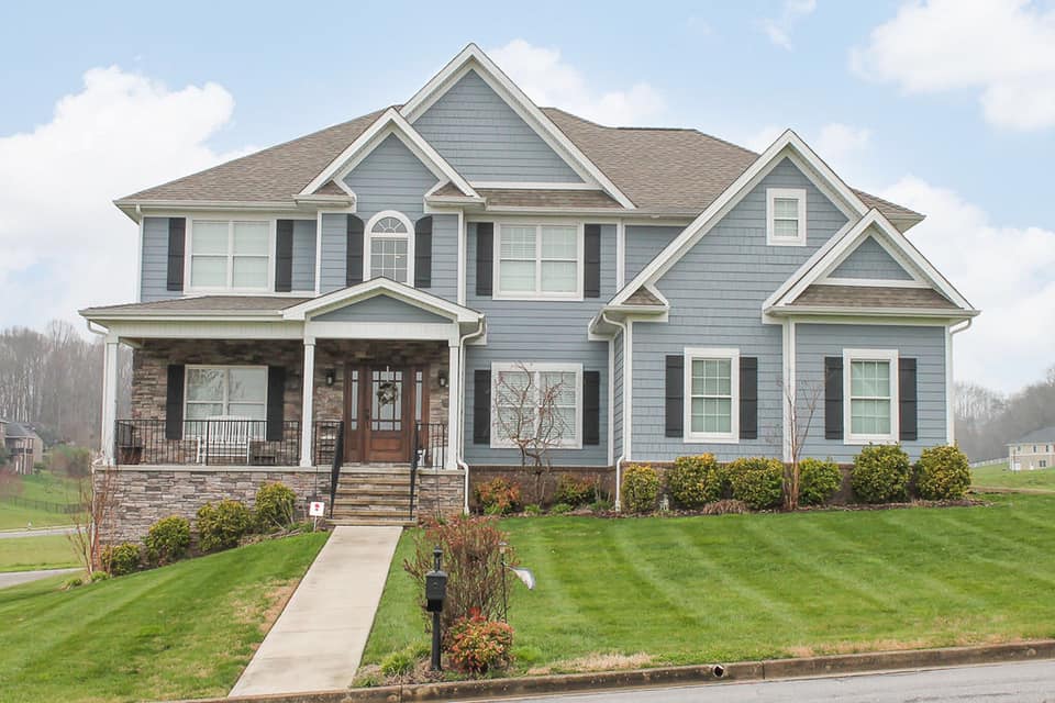 A large, two-story house with a light blue exterior, front porch, and landscaped lawn under a cloudy sky.