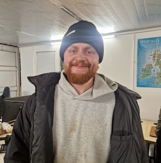 A man with a beard and a hat stands smiling indoors, dressed warmly in a jacket and hoodie.