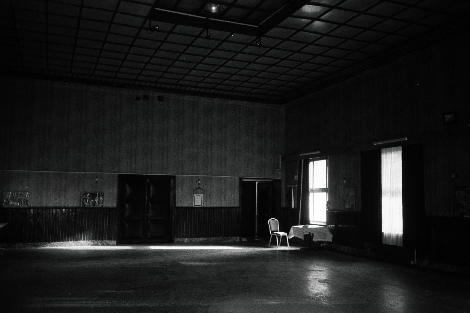 A dimly lit, empty room with a solitary chair positioned near a small table by two windows, casting soft natural light across the bare floor, creating an atmosphere of solitude and quiet reflection.