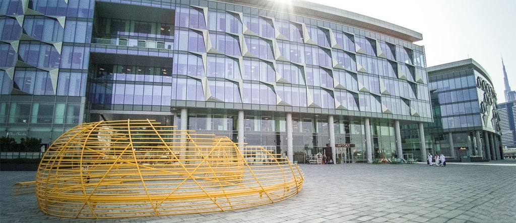 Modern glass building in Al Jaddaf with yellow sculpture, Art Cafe signage, and people in traditional attire.