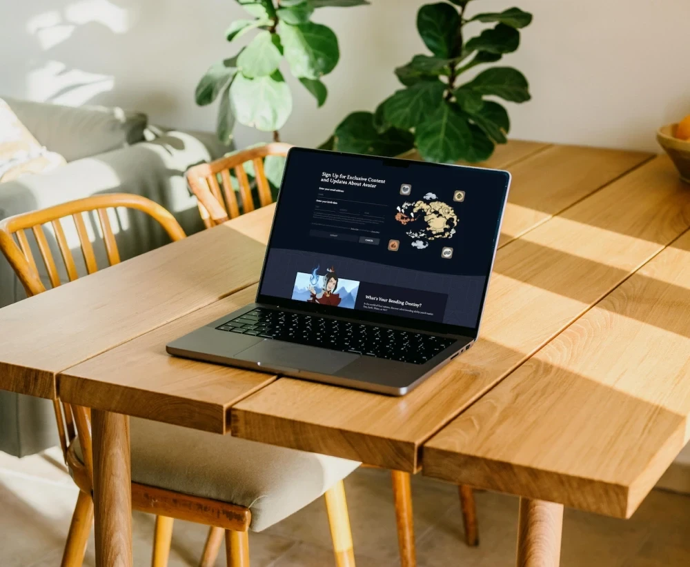 Fluid web design of a newsletter sign-up form and illustrated character card shown on a laptop in a sunlit room with wooden table and plants.