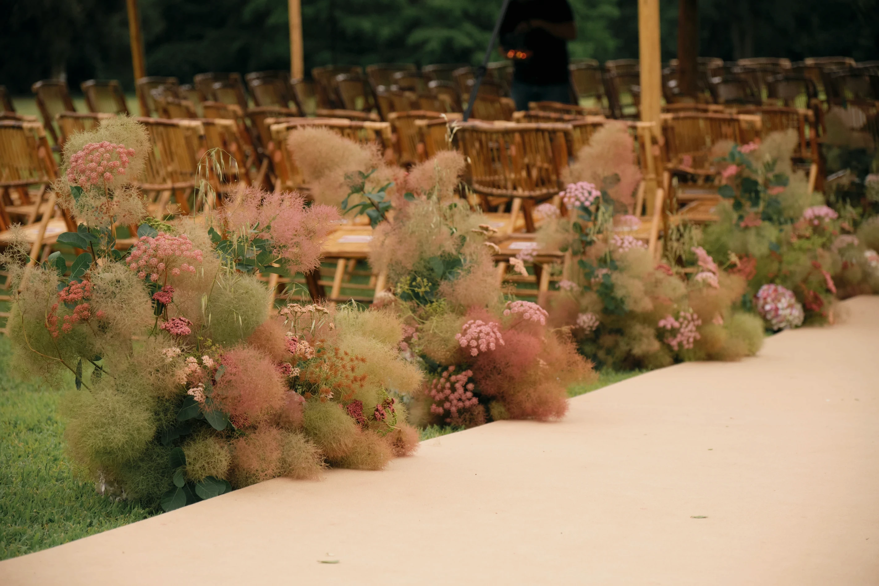 Camino de ceremonia con arbustos de smoke bush rosado y flores silvestres frente a hileras de sillas de caña.