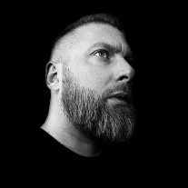 Young man in a dark suit jacket and cream sweater vest over a blue shirt with a patterned tie. Professional and confident expression, neutral background.