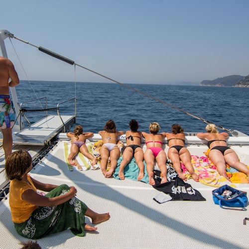 Des personnes se détendent et prennent un bain de soleil sur le pont d'un bateau en mer, avec la côte et les collines visibles au loin.
