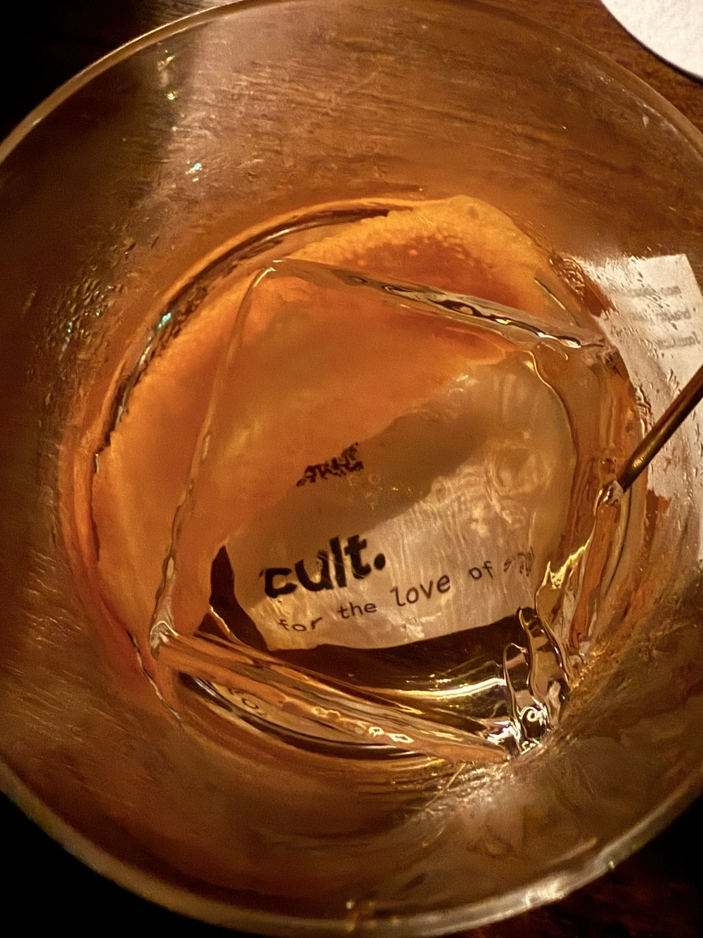 a business card for cultsool.com is visible through an ice cube at the bottom of a cocktail glass; an orange rind curves around the ice cube