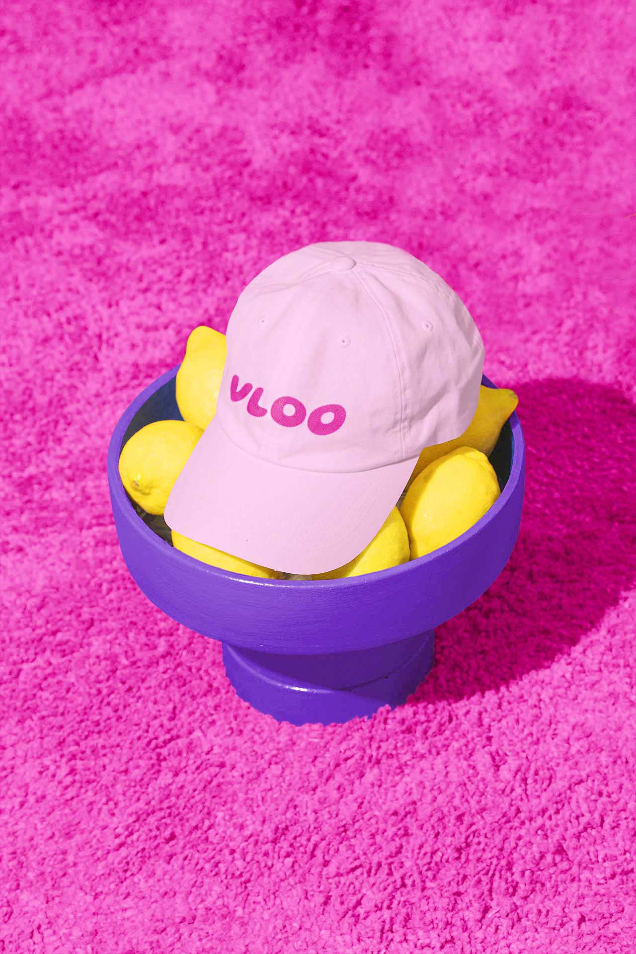 A pink VLOO hat with the logo, sitting on a purple bowl of lemons on a plush hot pink carpet.