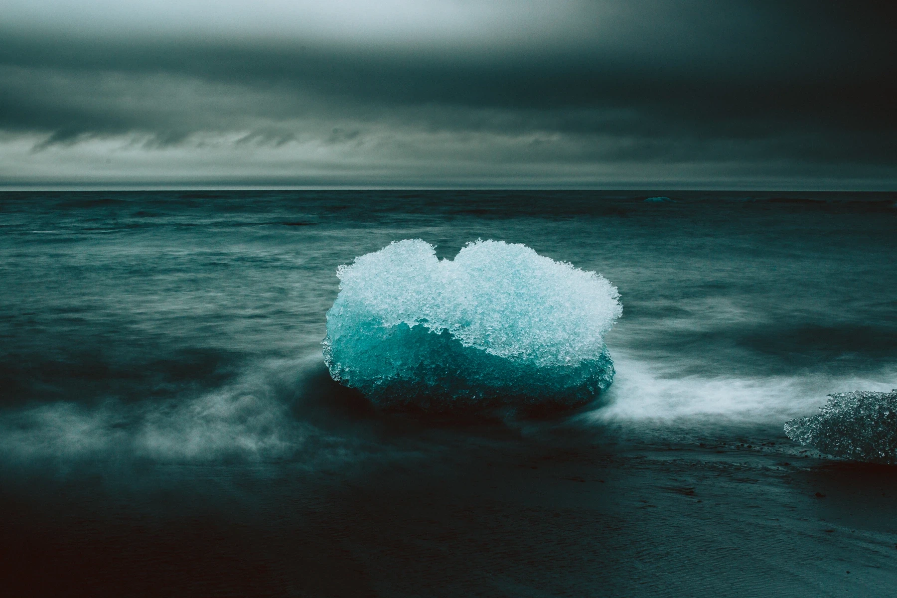 A small iceberg on black sand with surf washing around it under grey sky