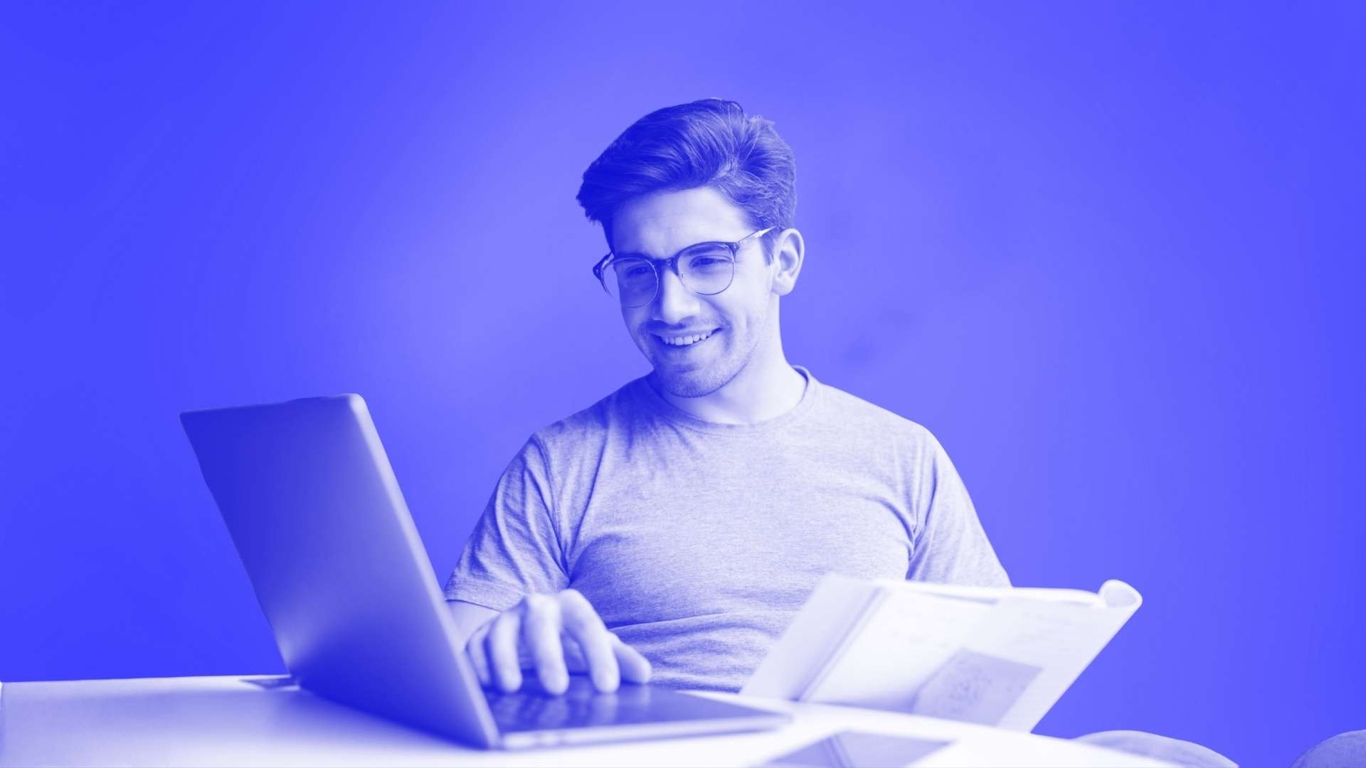 A smiling man sat at his desk with one hand on his laptop and another one holding a document