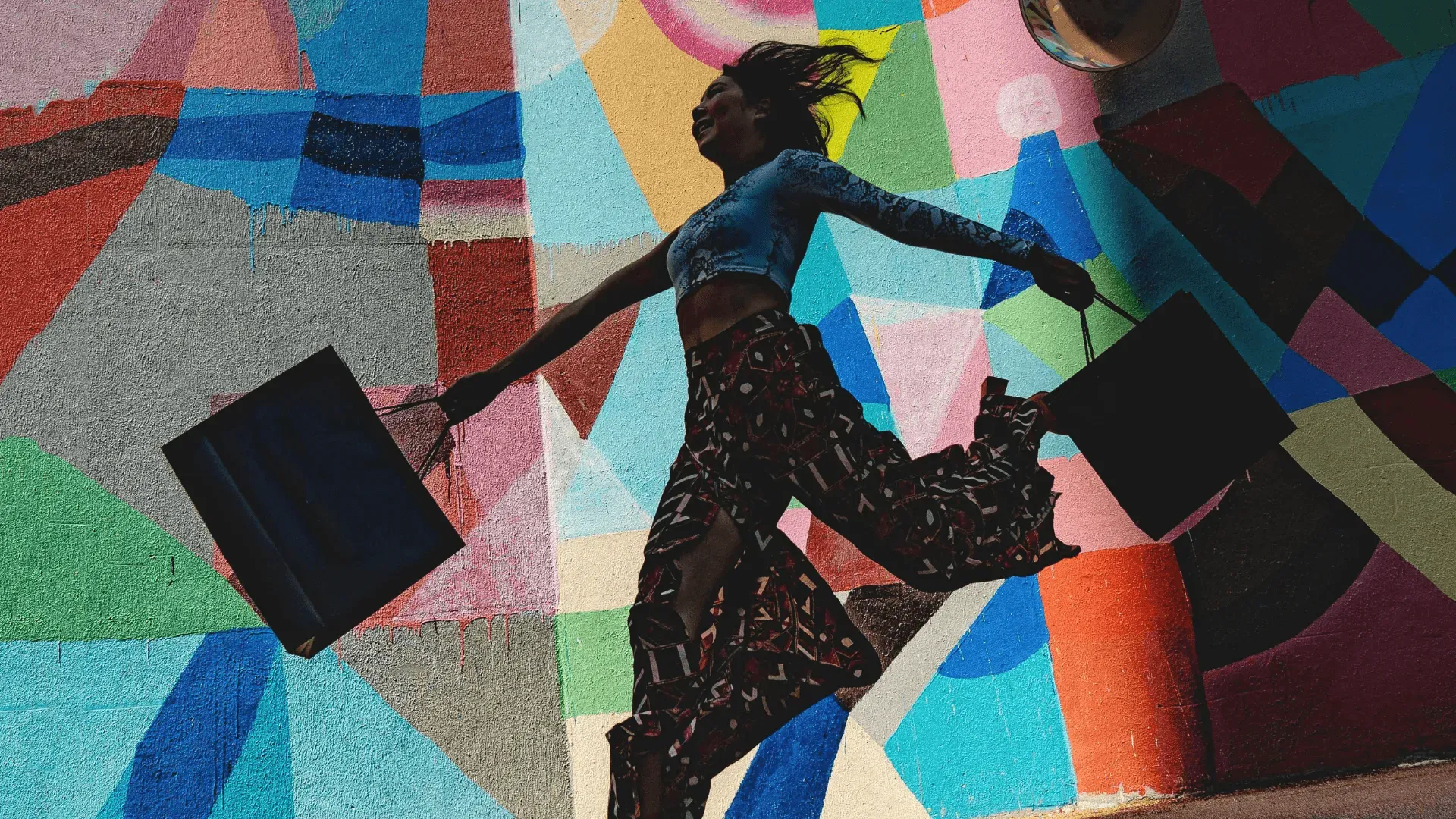 A woman leaping with arms outflung, holding shopping bags, in front of a brightly coloured wall