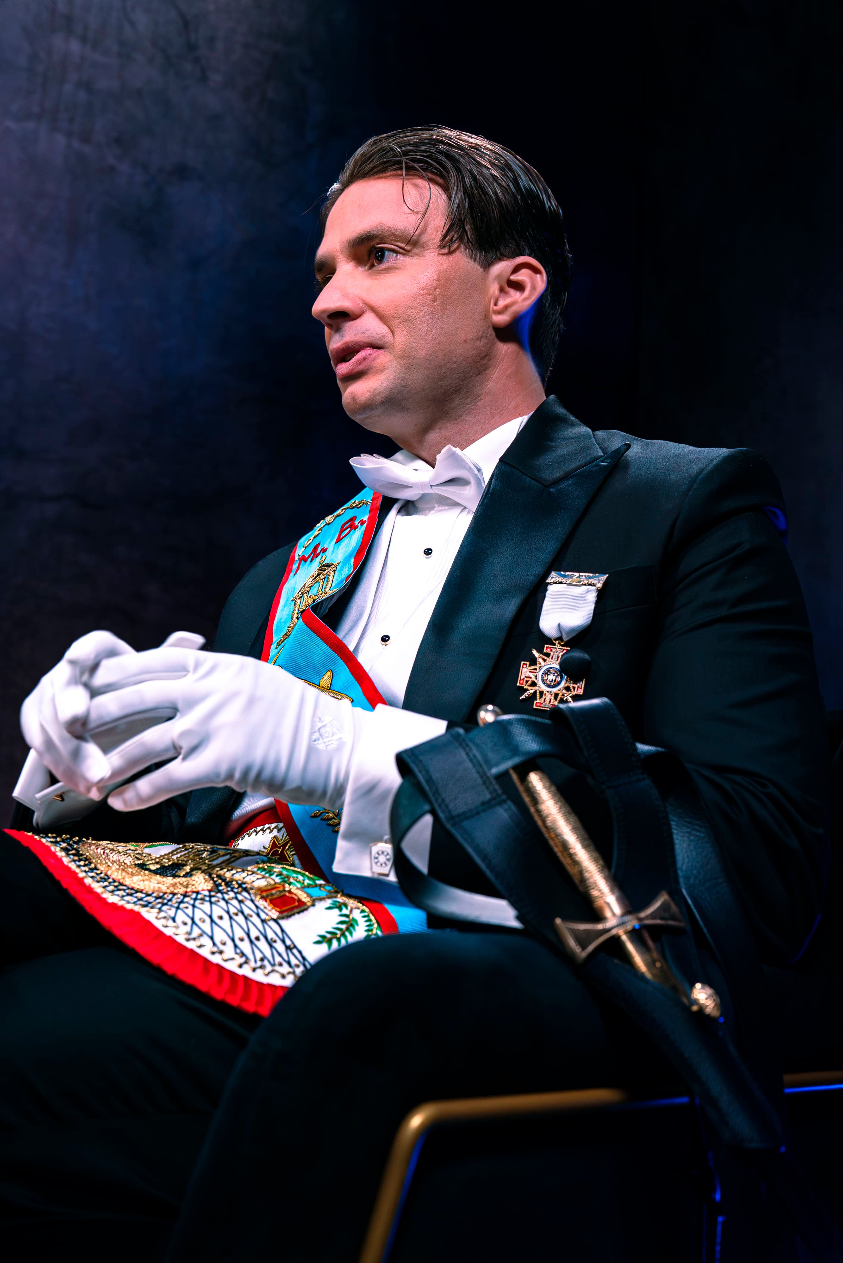 Photographie de portrait professionnelle d'un homme en tenue de Franc-Maçonnerie (smoking, gants blancs, écharpe d'officier colorée) interviewé dans le studio Legend. L'homme est assis, le regard dirigé hors champ, dans une posture d'écoute attentive. Une épée cérémonielle dans son fourreau est posée sur ses genoux. Le costume et les insignes sont très détaillés. L'arrière-plan est sombre, éclairé par des touches de lumière bleue. Ce portrait, optimisé par Nux-Studio, illustre la production photo pour des interviews formelles, des sujets d'enquête et de société, mettant en évidence l'éclairage de studio et la direction artistique.