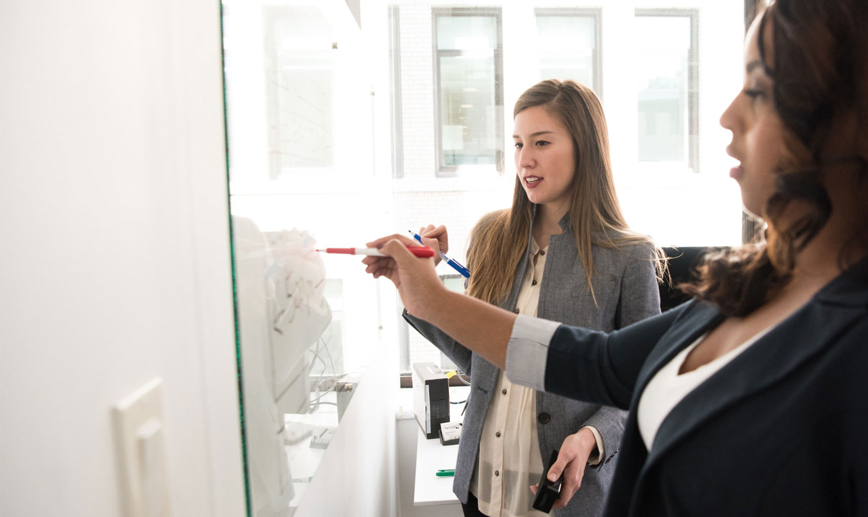 Two women in a meeting; one is writing on a whiteboard while the other observes attentively.