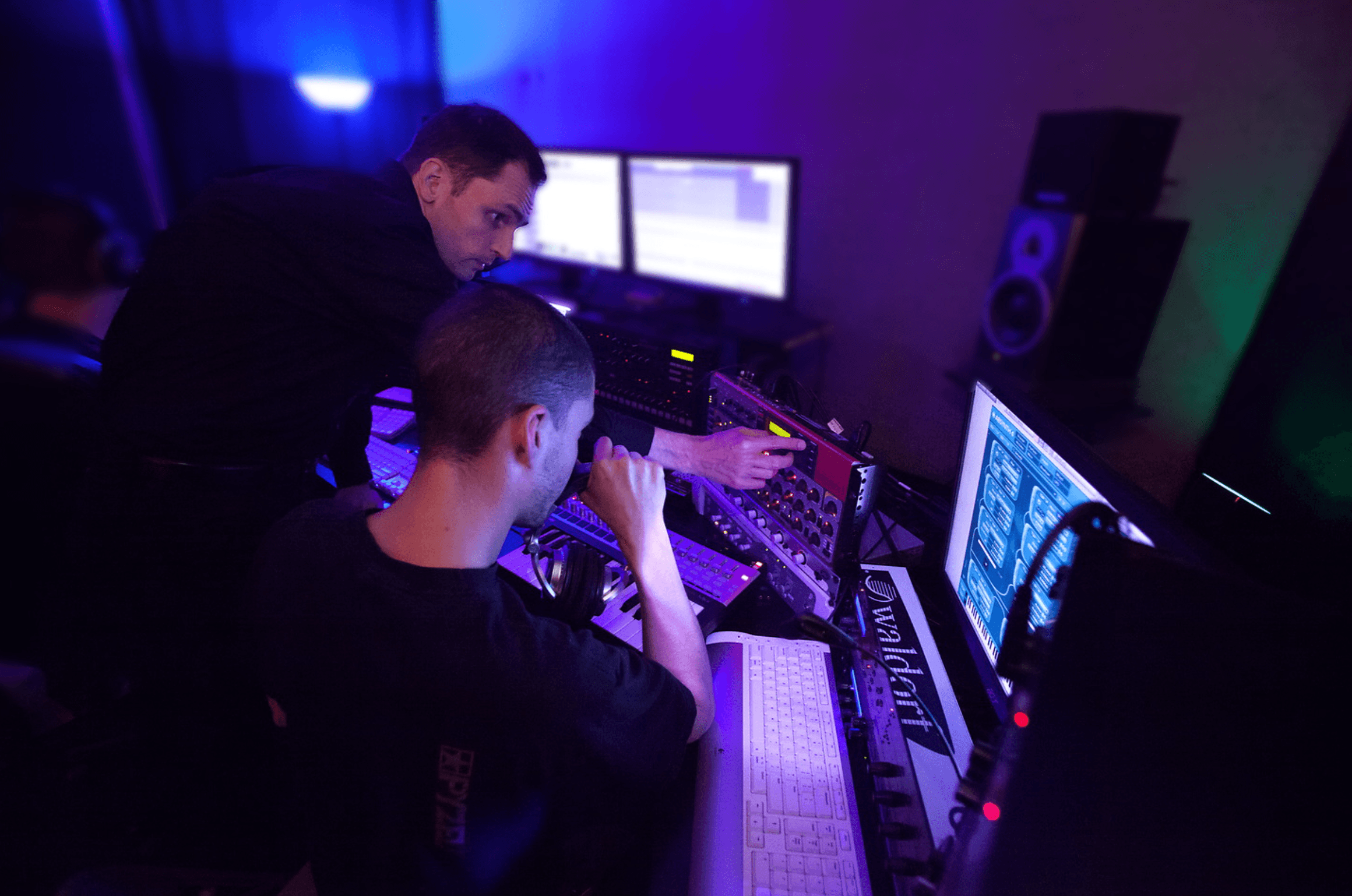 In a dimly lit music studio, two people are focused on mixing soundtracks at a workstation, surrounded by multiple screens and audio equipment, highlighting a collaborative process in audio production.