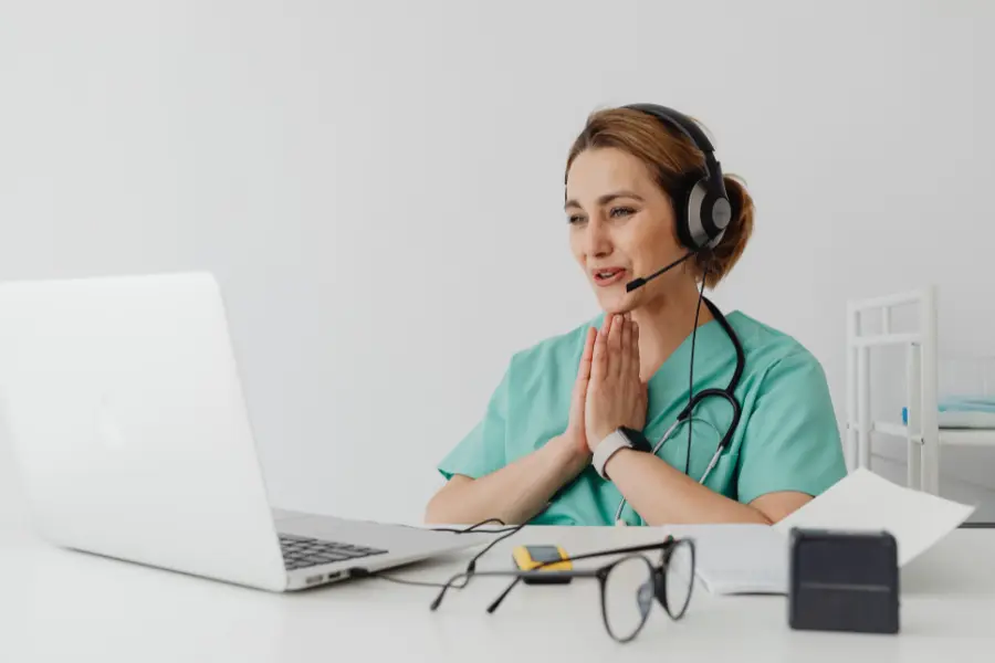 AI admin assistant for healthcare shown as a nurse in green scrubs and a headset speaks attentively into a laptop during a virtual consultation at her desk.