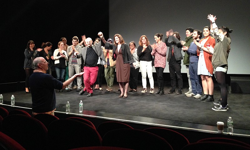 A photo of a group of people standing on a stage, applauding and holding each others hands up.
