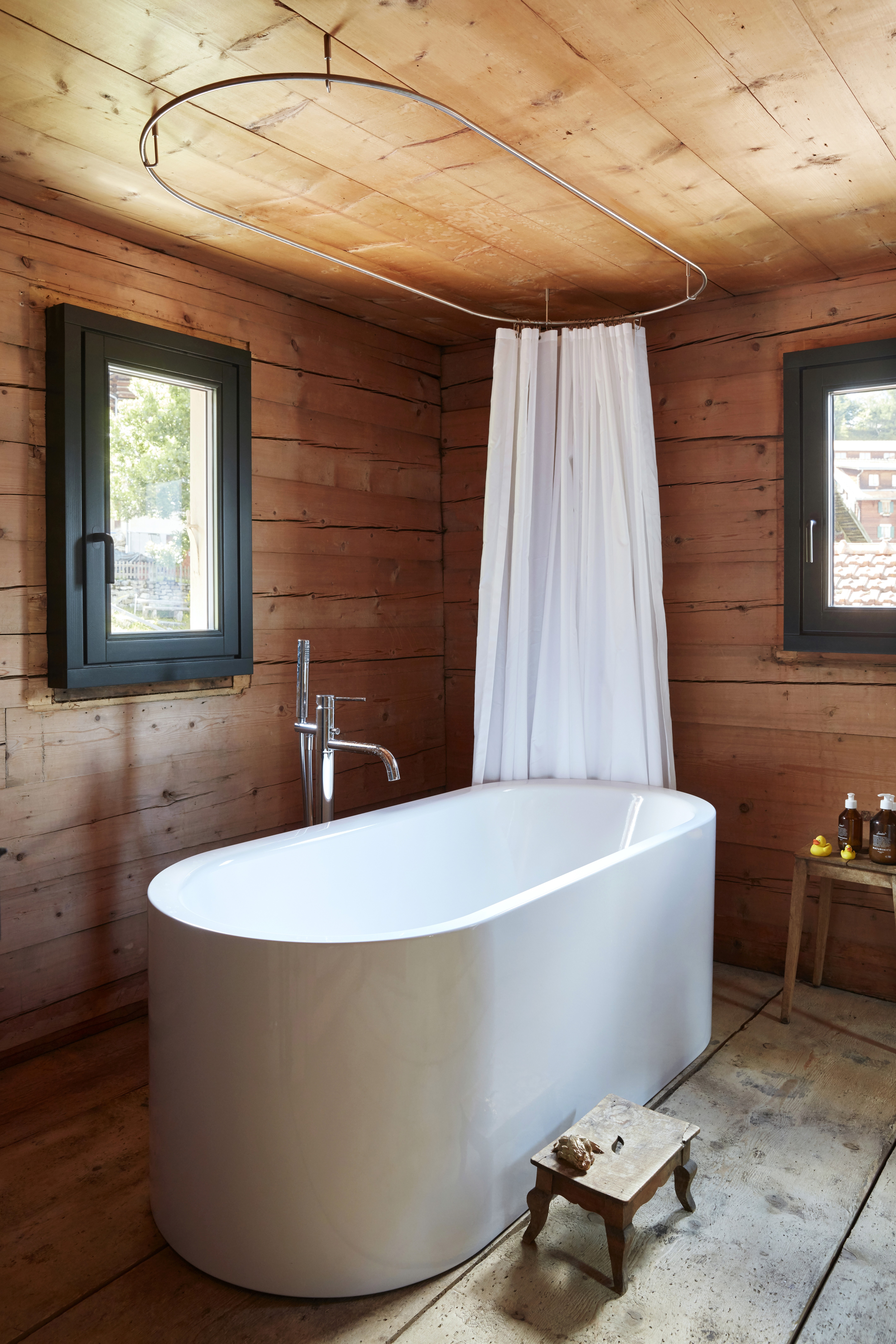 A free standing white bathtub in a wooden bathroom with a white curtain.