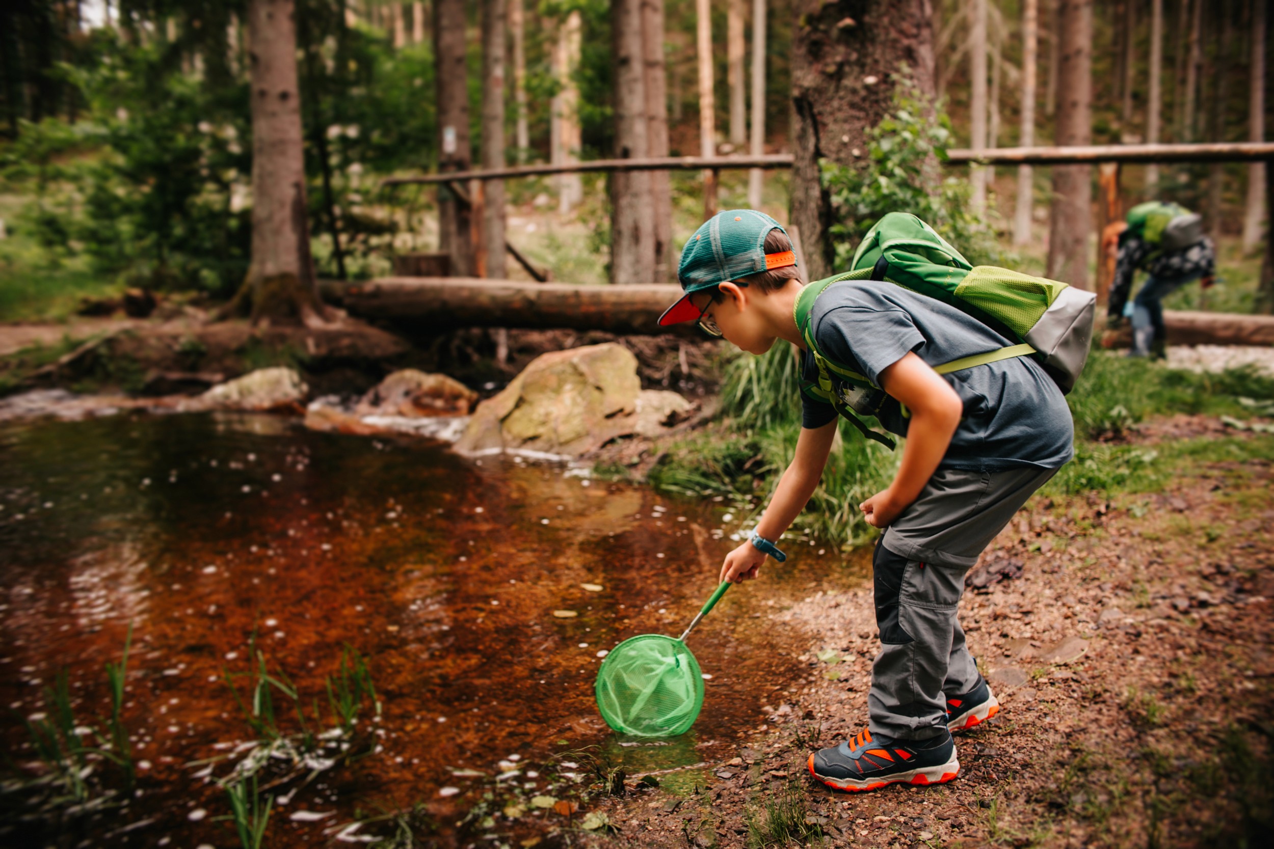 Kind mit grünem Rucksack und Kecher am Bach