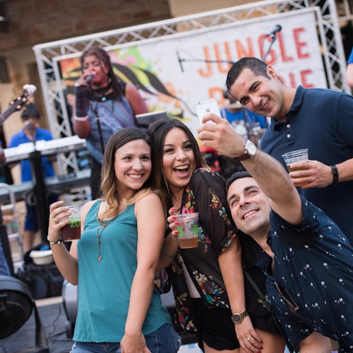 Four people are smiling and taking a selfie at an outdoor event, holding drinks, with a singer performing in the background.