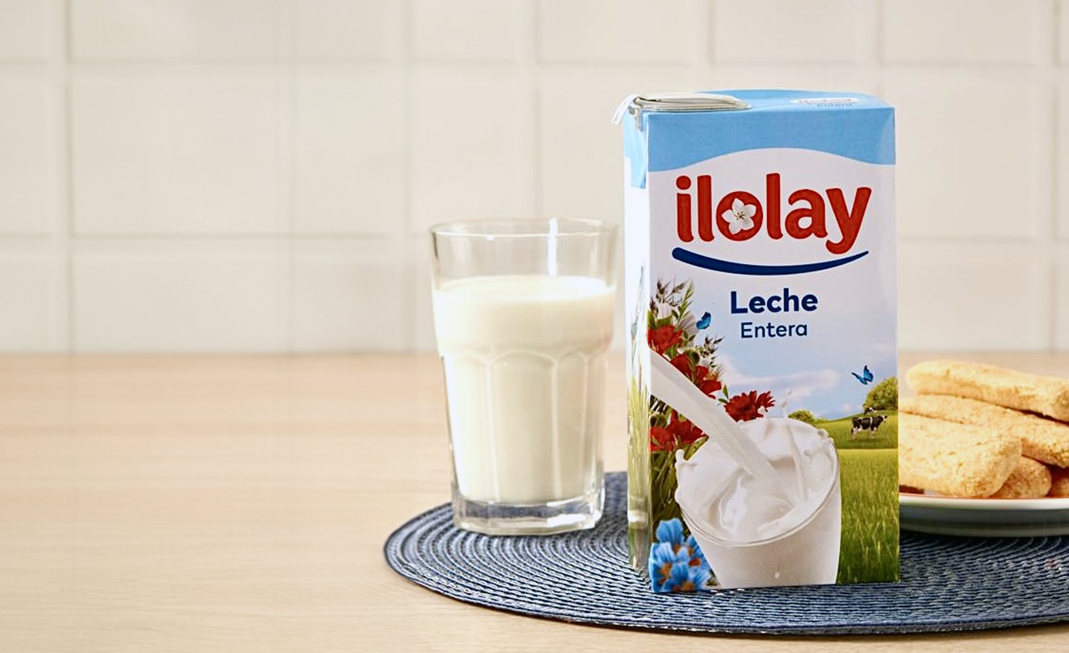 A carton of milk, a glass of milk, and a biscuit on a plate against a light background.
